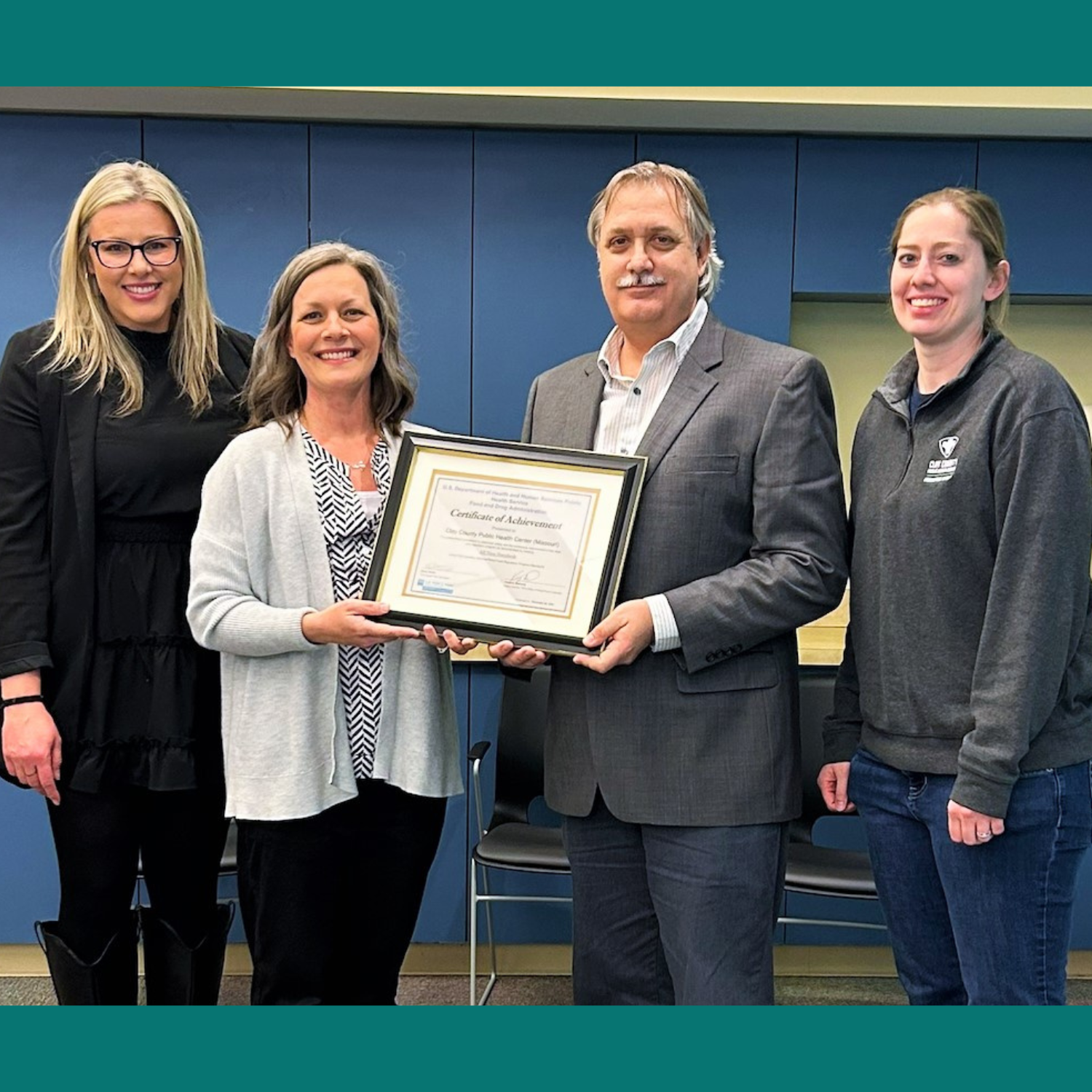 Four adults stand in a row, smiling and holding a framed certificate