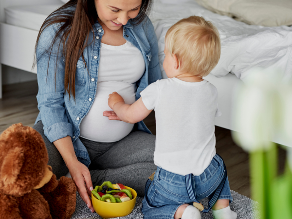 Pregnant woman sits on bedroom floor with a bowl of fruit and a young boy