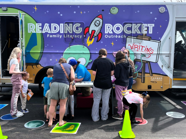 People standing at a table outside in front of a library truck called the Reading Rocket